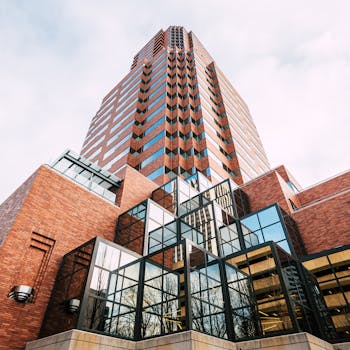 From below of multistory contemporary multistage building with geometric facade made of stone bricks and metal panels with trees reflecting in glass windows under cloudy sky on sunny day